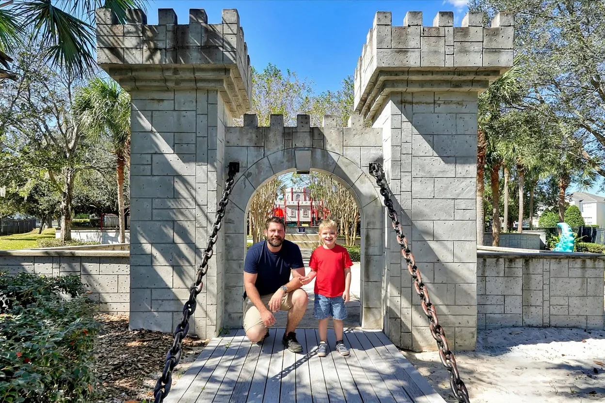 A dad crouching beside his young son, who stands up tall in a red t-shirt, in front of a stone castle archway