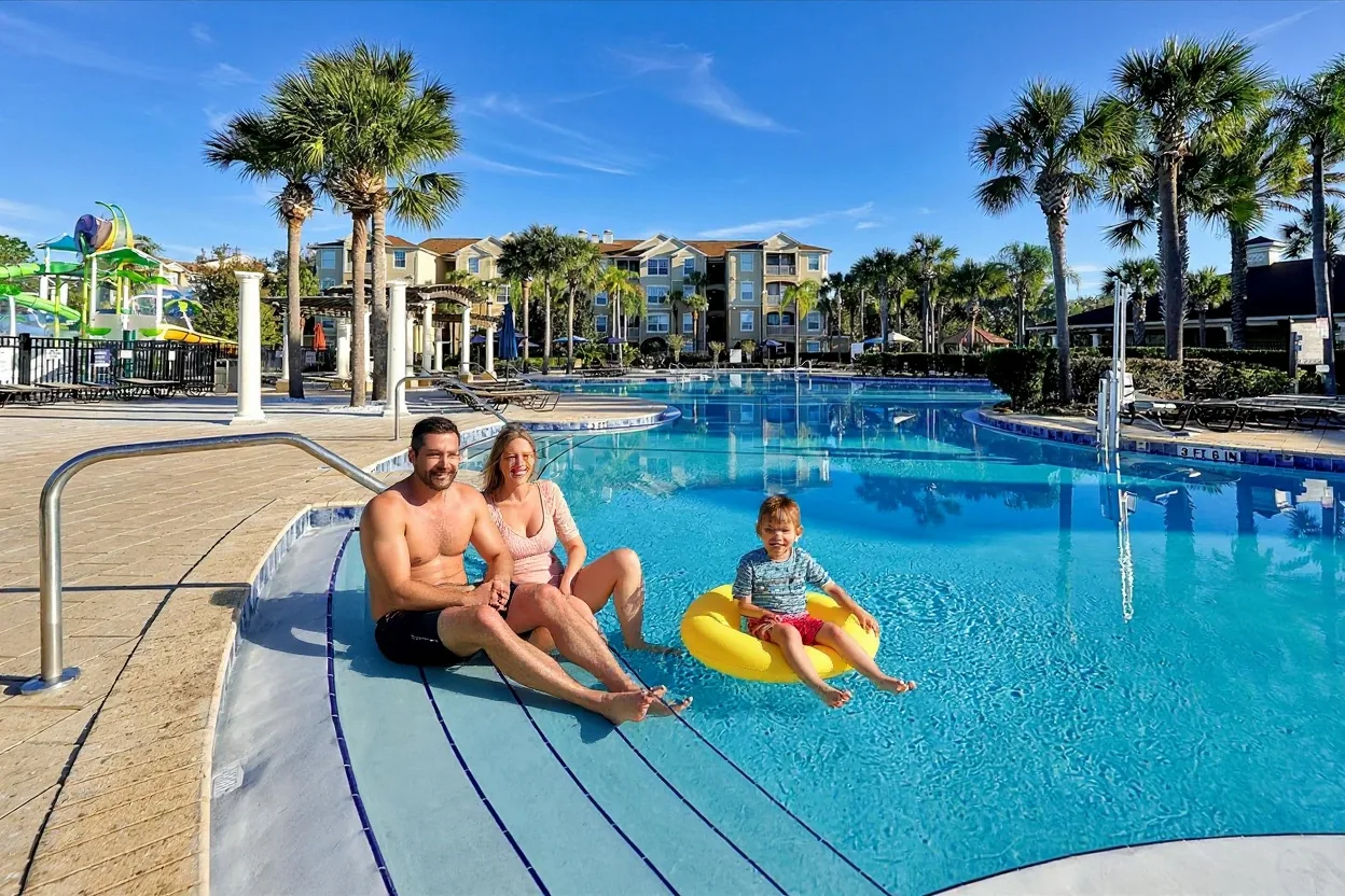 An ordinary family relaxing on a rest day: parents sitting on the pool steps in plain swimwear and two kids in rash guards floating on bright noodles