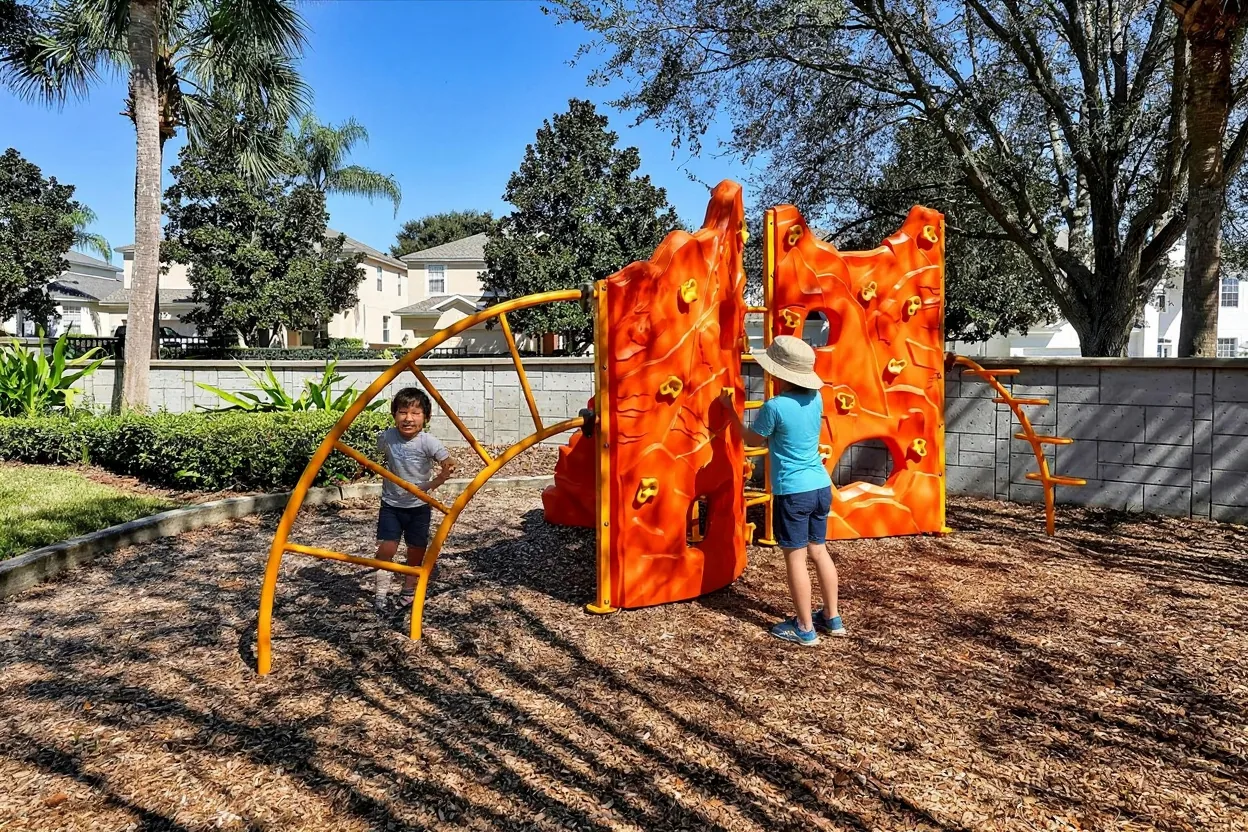 A 3-year-old in a plain t-shirt and shorts climbing the low holds on the existing climbing wall with a mom in a sun hat spotting from behind, both