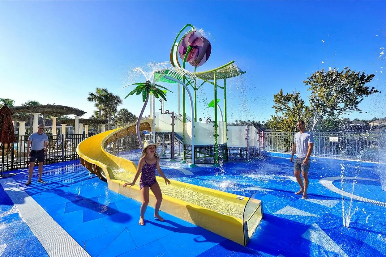 A 4-year-old girl in a one-piece swimsuit and sun hat laughing under the existing water spray, with an ordinary dad in a plain t-shirt watching nearby