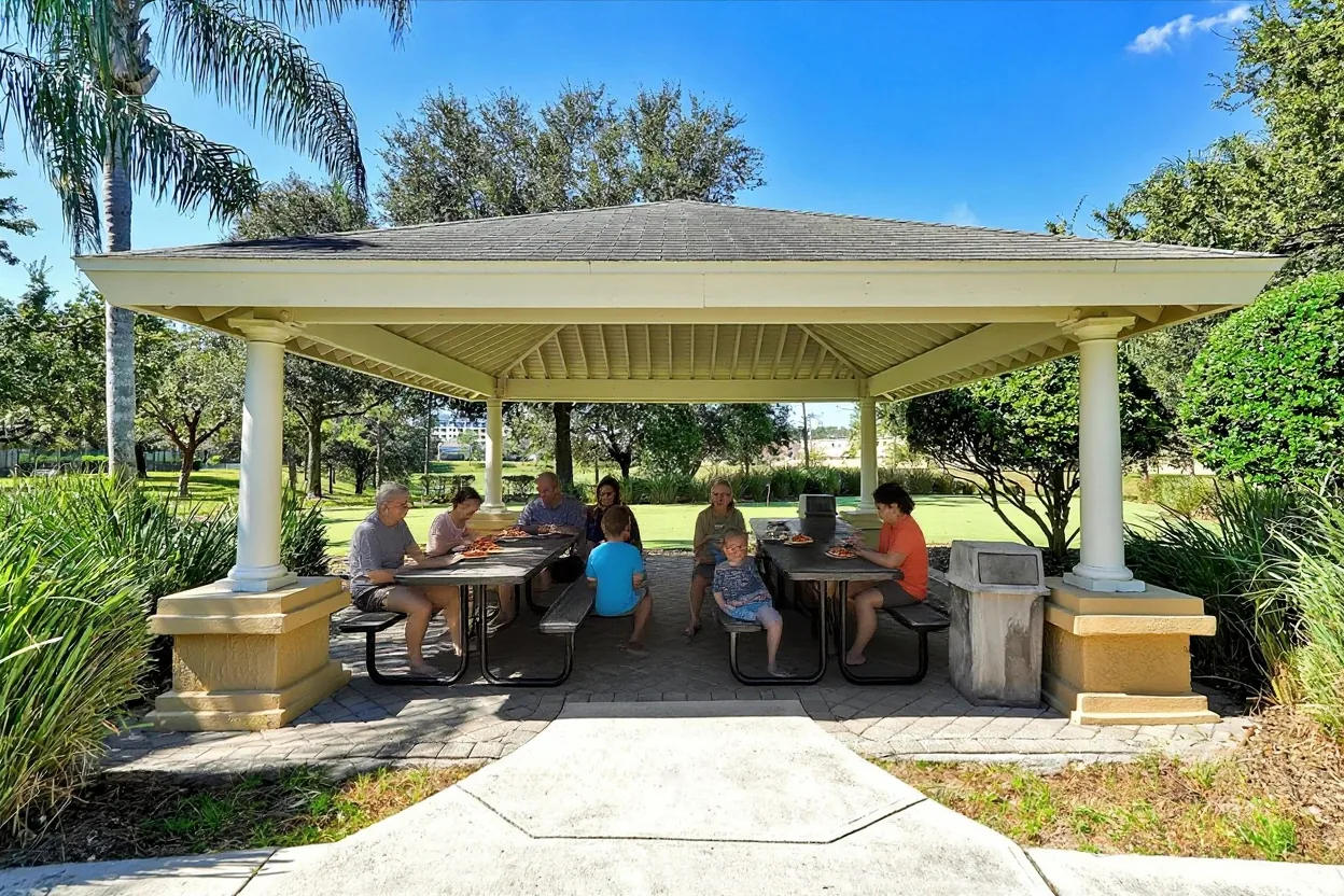 A big extended family of ten eating pizza at the existing picnic tables under the pavilion roof: grandparents, parents and kids of different ages in