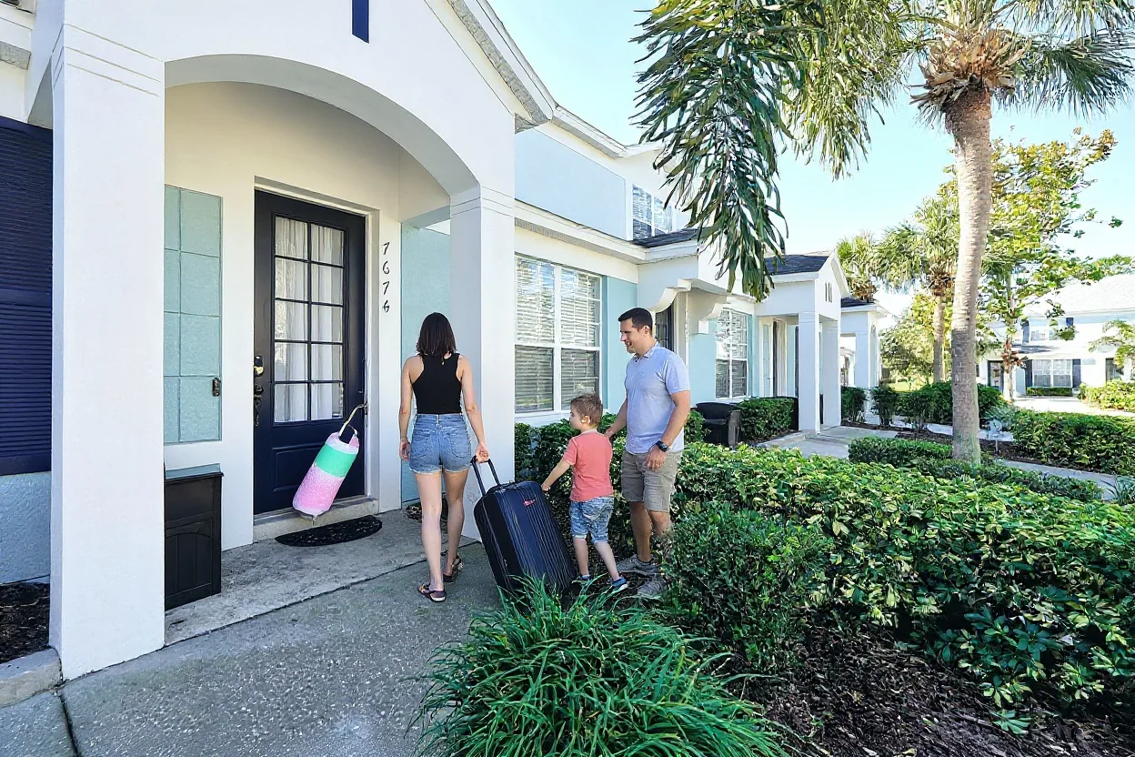 An ordinary family arriving at the townhouse front door: a mom unlocking the door with a keypad, a dad with two rolling suitcases and a kid